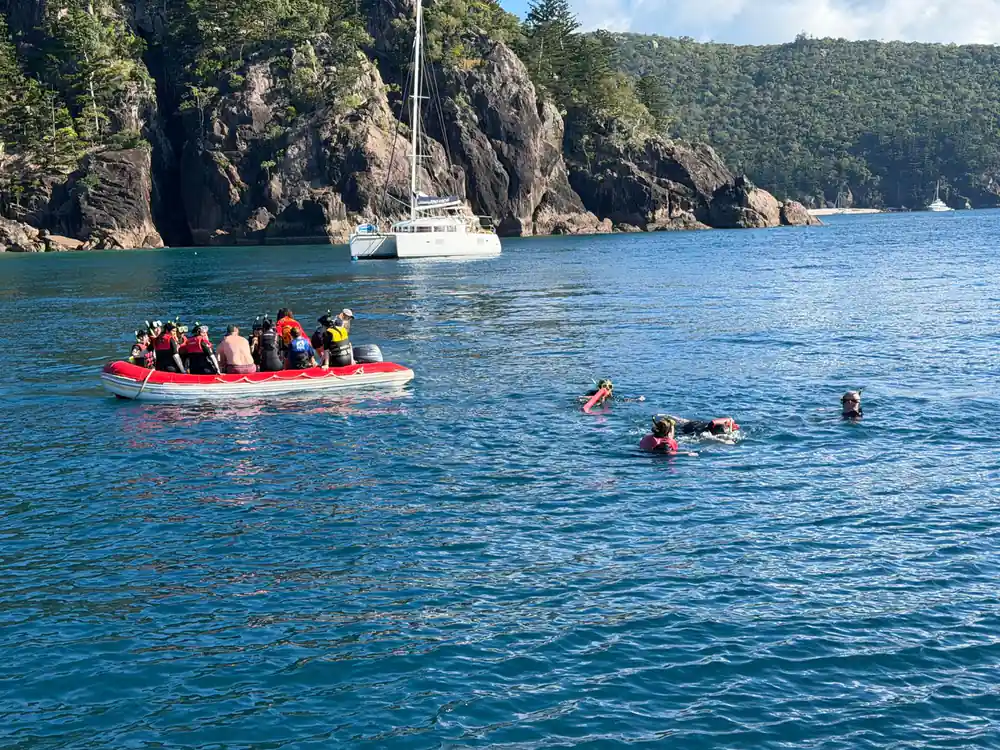 Tourists in red boat snorkeling near rocky cliff and sailboat